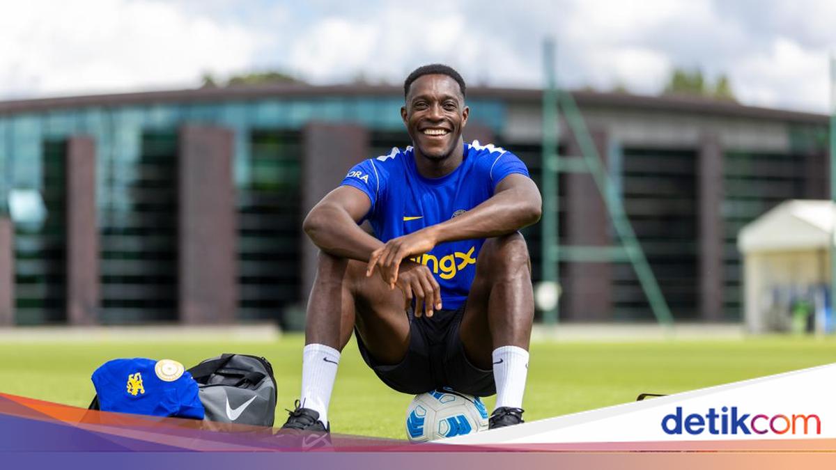 Seorang pemain sepak bola berbaju biru tersenyum sambil duduk di atas bola di lapangan latihan.