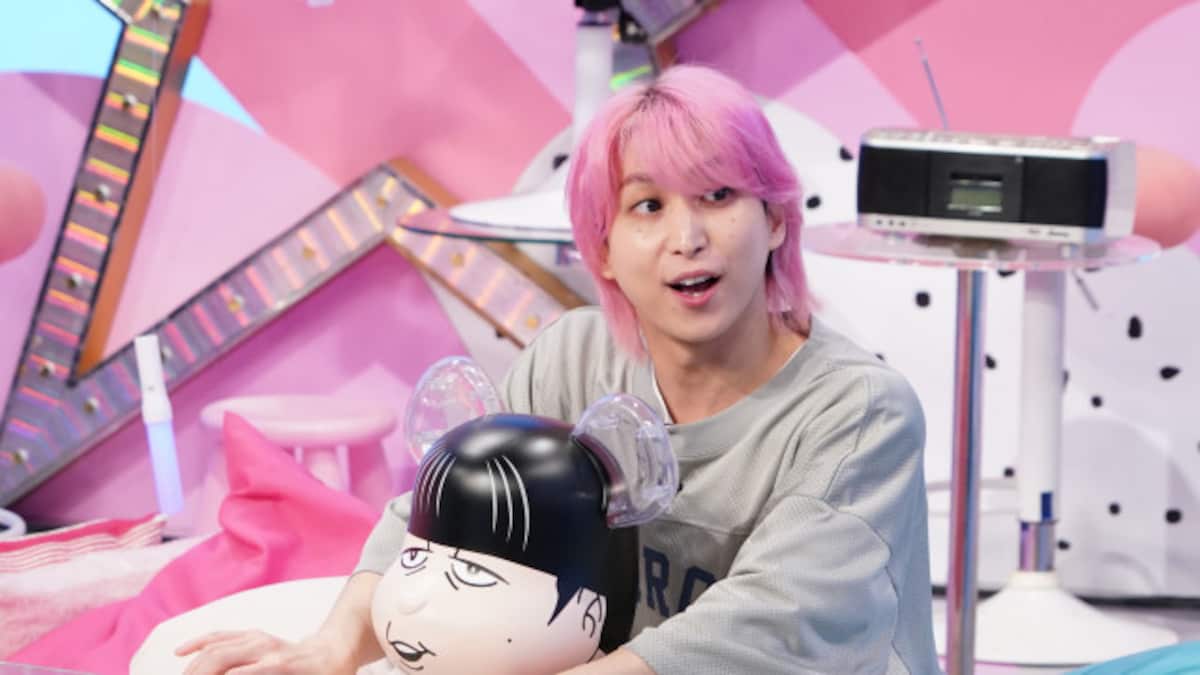 ピンク色の髪の出演者が、カラフルなテレビ番組のセットでキャラクターの小道具に手を添えている。